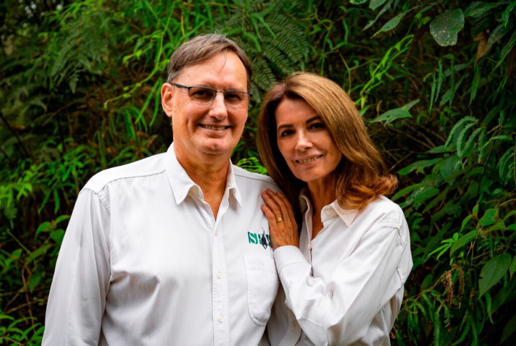 Imagem de Clóvis Borges e Mônica Borges se abraçando e sorrindo para a foto. Ambos são brancos e estão usando camisas sociais brancas. Clóvis usa óculos e tem cabelo liso castanho curto, Mônica tem cabelo liso na altura dos ombros.